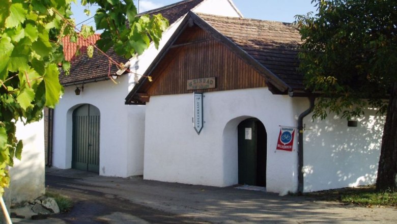 Weinkeller, © Fam. Döller Weißes Gebäude mit Holzdach, Weinreben im Vordergrund.