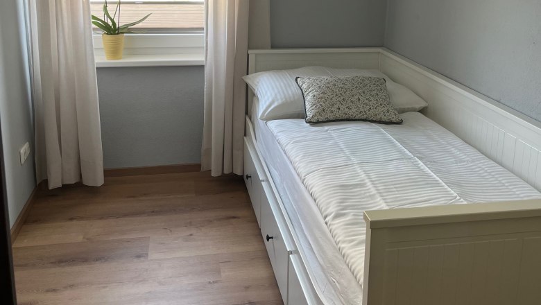 Schlafzimmer, © Jochen Maurer Kleines Schlafzimmer mit Einzelbett, weißen Vorhängen und einer Pflanze auf der Fensterbank.