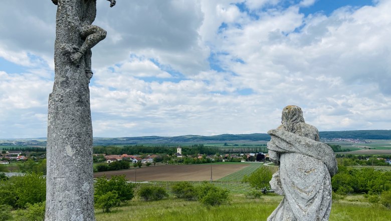 Riede Kalvarienberg, © Weinstraße Weinviertel Zwei Steinskulpturen auf einem Hügel mit Blick auf eine ländliche Landschaft und ein Dorf im Hintergrund.