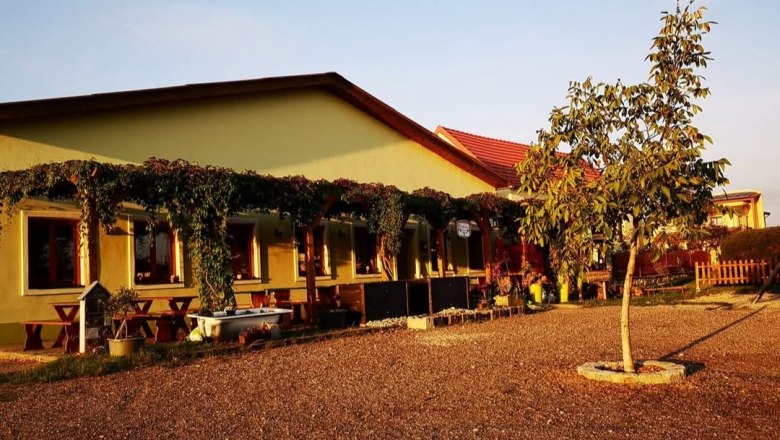 Dorfheurigen, © M. Vittek Gelbes Gebäude mit Terrasse, Weinreben und Baum im Vordergrund bei Sonnenuntergang.