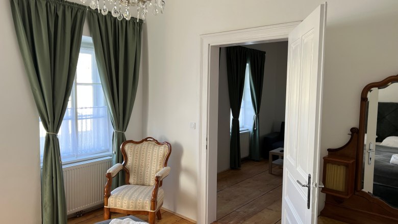 Schlafzimmer, © Balcar Ein elegantes Schlafzimmer mit einem antiken Sessel, grünen Vorhängen und einem Kronleuchter. Eine offene Tür führt in einen angrenzenden Raum.