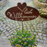 Herzlich Willkommen, © Familie Fleischmann Metallschild mit 'Herzlich Willkommen' über einem Topf mit gelb-violetten Blumen auf einem gepflasterten Weg.