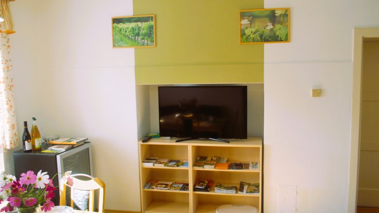 Weinbauernhof Löscher, Esszimmer, © Christina Löscher Ein Wohnzimmer mit einem Fernseher auf einem Regal, umgeben von Broschüren. An der Wand hängen zwei Bilder von Weinbergen. Ein Tisch mit Blumen und Geschirr steht links.