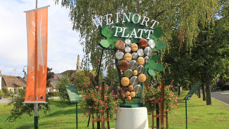 Größte Weintraube in NÖ, © Dorferneuerungsverein Platt Willkommensschild in Platt mit Weintrauben-Dekoration und Kirche im Hintergrund.