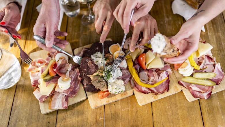 Heurigenjause, © Weinviertel Tourismus GmbH / Herbst Mehrere Hände greifen nach Wurst- und Käseplatten auf einem Holztisch.