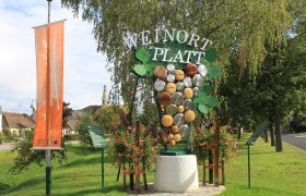 Größte Weintraube in NÖ, © Dorferneuerungsverein Platt Willkommensschild in Platt mit Weintrauben-Dekoration und Kirche im Hintergrund.