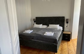 Schlafzimmer, © Balcar Ein gemütliches Schlafzimmer mit einem großen Bett, grauer Bettwäsche und weißen Handtüchern. Ein Kronleuchter hängt von der Decke.