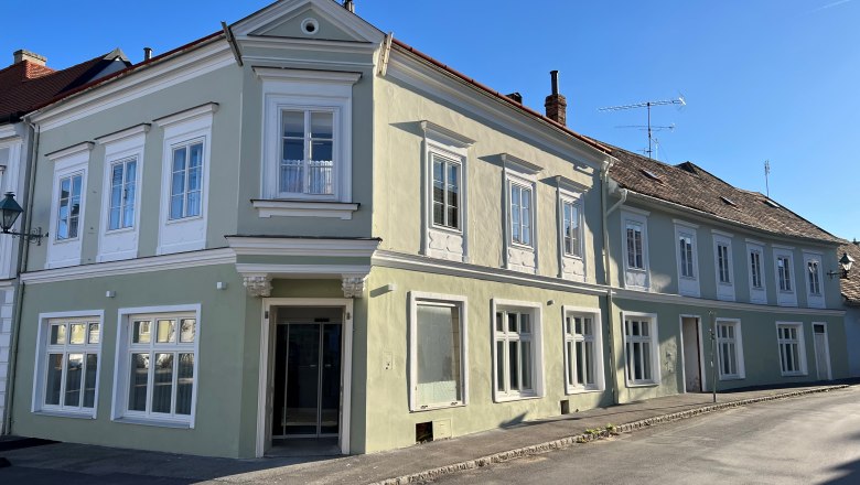 Außenansicht, © Balcar Ein zweistöckiges, pastellgrünes Gebäude mit weißen Fensterrahmen an einer Straßenecke bei klarem Himmel.