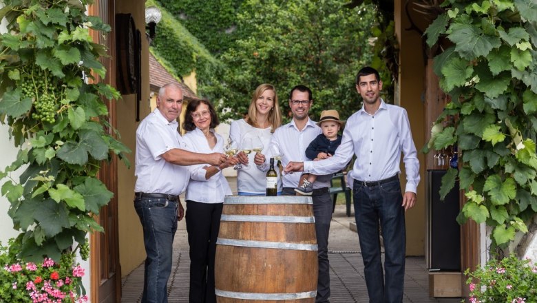 Familie Pöcher, © Andreas Schweighofer Eine Familie steht um ein Weinfass, umgeben von Weinreben, und stößt mit Weingläsern an.