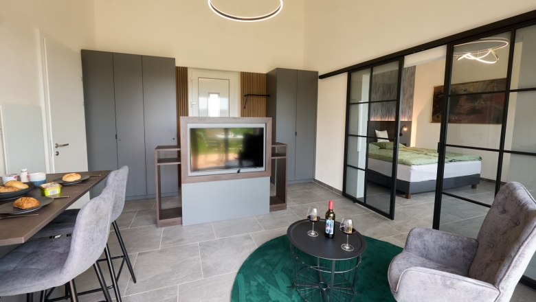 Apartments Vineyard19, © Apartments Vineyard19 Modernes Apartment mit Esstisch, Fernseher, grauen Schränken und Glas-Schiebetüren zum Schlafzimmer.