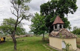Heiliger Stein, © Weinstraße Weinviertel Kleine Kapelle mit rotem Ziegeldach in einer grünen Landschaft, umgeben von Bäumen und Bänken.