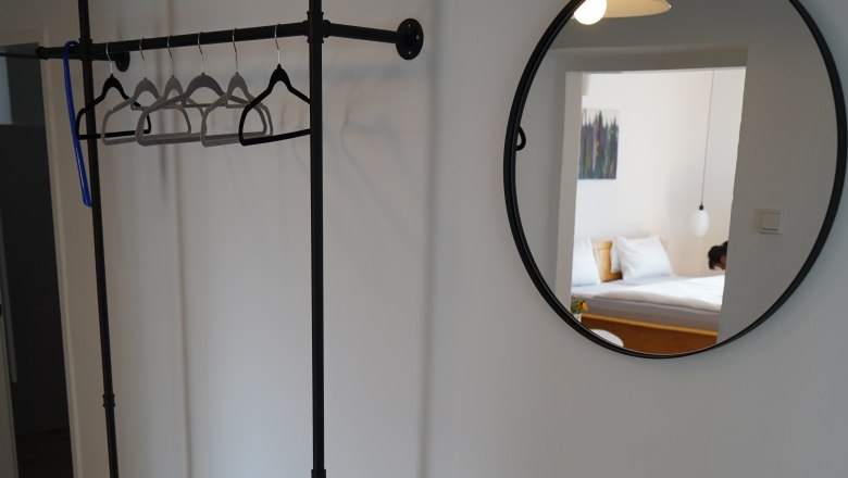 Landhaus Krell, © Weingut Krell Innenansicht eines Zimmers mit Kleiderständer, rundem Spiegel und kleinem Tisch mit Pflanze und Vase.