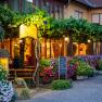 Gastgarten, © Martin Lifka Photography Ein gemütlicher Gastgarten mit blühenden Pflanzen und Weinreben, beleuchtet von warmem Licht. Menschen sitzen an Holztischen unter einem Laubengang.
