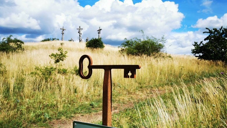 Kalvarienberg Schrattenthal, © Weinstraße Weinviertel Ein Fotopunkt-Schild vor einer Wiese mit drei Kreuzen im Hintergrund unter blauem Himmel mit Wolken.