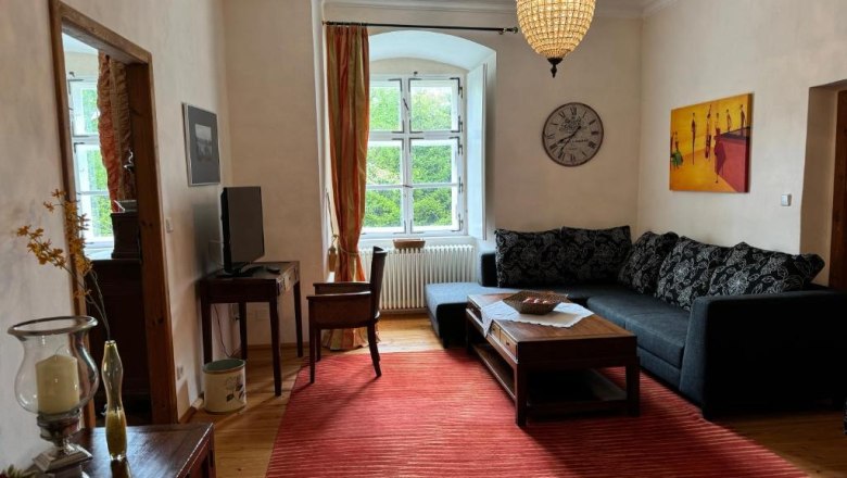 Ferienwohnung, © Andrea Schubert Gemütliches Wohnzimmer mit Sofa, rotem Teppich, Kronleuchter und großem Fenster.