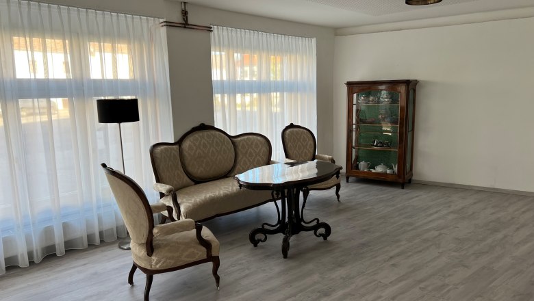 Eingangsbereich, © Balcar Ein eleganter Raum mit antiken Möbeln, darunter ein Sofa, ein Tisch und ein Schrank mit Geschirr. Große Fenster mit weißen Vorhängen lassen viel Licht herein.