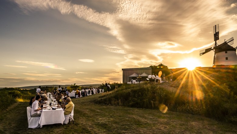 Tafeln im Weinviertel, © Weinviertel Tourismus GmbH / www.pov.at Menschen essen an langen Tischen im Freien bei Sonnenuntergang neben einer Windmühle.
