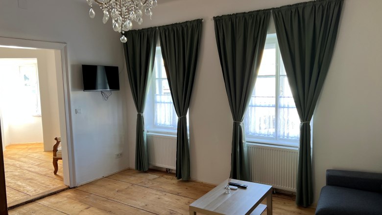 Gästezimmer, © Balcar Ein Wohnzimmer mit Holzboden, einem Kronleuchter, zwei Fenstern mit grünen Vorhängen, einem grauen Sofa und einem kleinen Holztisch.