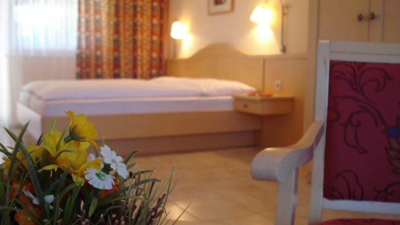 Gästezimmer, © Pension Rudi Ein gemütliches Gästezimmer mit einem Bett, einem Stuhl und einem Tisch mit Blumen.