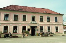 Ein zweistöckiges, rosa Gebäude mit einem roten Ziegeldach und mehreren Fenstern. Vor dem Gebäude stehen Pflanzen und ein Motorrad.