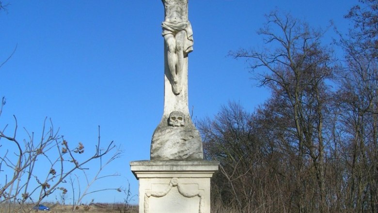 Steinkreuz mit Jesusfigur in einer verwilderten Landschaft unter blauem Himmel.