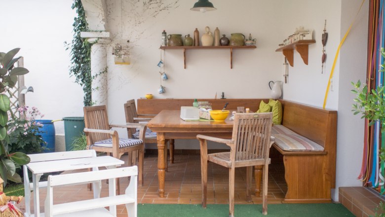 Gem&uuml;tliche Terrasse mit Holzm&ouml;beln, Eckbank, Tisch und Dekorationen. Pflanzen und Regale mit Keramik im Hintergrund.