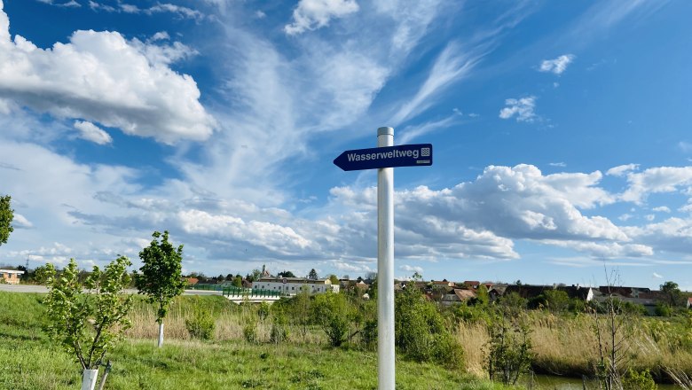 Ein Wegweiser mit der Aufschrift 'Wasserweltweg' steht auf einer grünen Wiese unter einem blauen Himmel mit weißen Wolken.