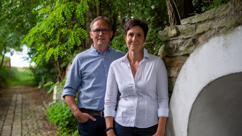 Ein Mann und eine Frau stehen nebeneinander auf einem gepflasterten Weg im Freien, umgeben von Bäumen und einer Steinmauer.