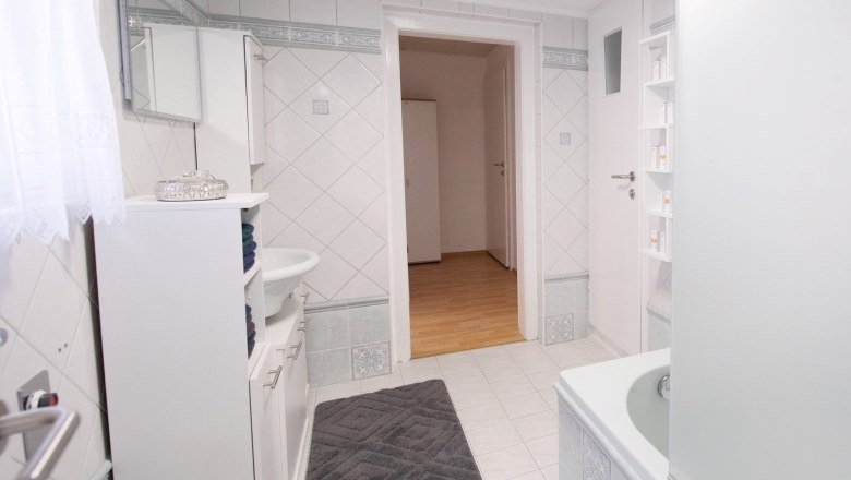 Modernes Badezimmer mit weißen Fliesen, Badewanne, Waschbecken und Schrank. Tür führt zu einem Flur mit Holzboden.