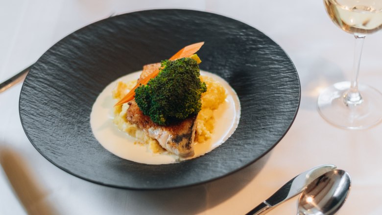 Ein Teller mit einem Gericht aus Fisch, Brokkoli und Kartoffelp&uuml;ree auf einem gedeckten Tisch mit Besteck und einem Glas Wei&szlig;wein.