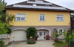 Gelbes Haus mit Solarpanels auf dem Dach, Garage und Eingang mit Blumen.
