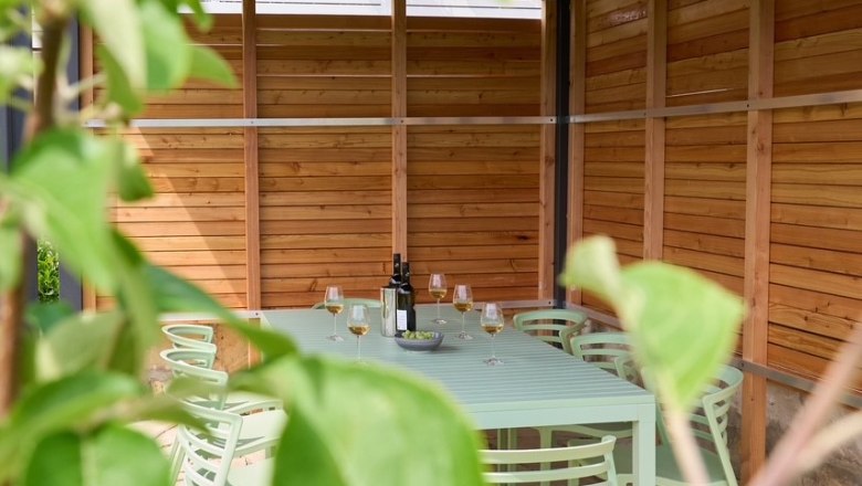 Überdachte Terrasse mit Tisch und Stühlen, umgeben von Holzverkleidung und Pflanzen im Vordergrund.