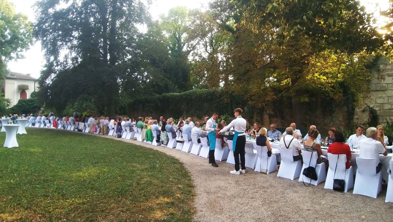 Langes Bankett im Freien mit vielen G&auml;sten an wei&szlig; gedeckten Tischen, umgeben von B&auml;umen.