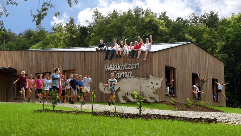 Eine Gruppe von Kindern und Erwachsenen sitzt auf einem Holzgeb&auml;ude mit der Aufschrift 'Wildkatzen Camp'. Einige Kinder stehen und springen davor.
