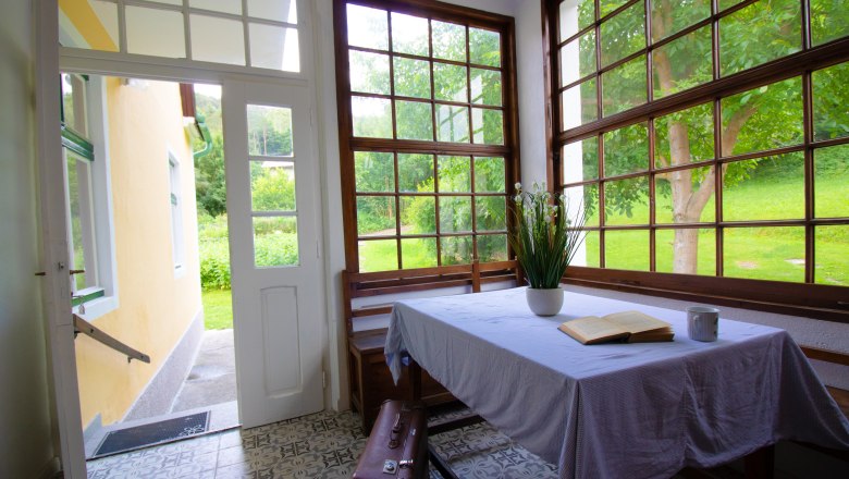 Innenansicht eines hellen Raumes mit Tisch, Buch, Tasse und Pflanzen, umgeben von gro&szlig;en Fenstern mit Blick auf einen Garten.