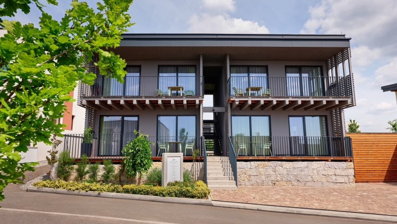 Moderne zweistöckige Apartmentanlage mit Balkonen und Treppenaufgang, umgeben von Pflanzen und Bäumen.