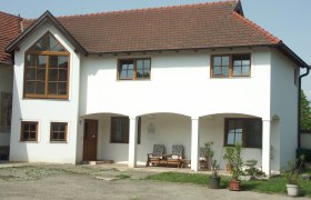 Ein zweist&ouml;ckiges wei&szlig;es Geb&auml;ude mit roten Dachziegeln und gro&szlig;en Fenstern. Vor dem Haus stehen Pflanzen in T&ouml;pfen.