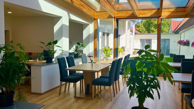 Ein heller Raum mit Glasdach, Holzboden, Pflanzen und Tischen mit blauen St&uuml;hlen.