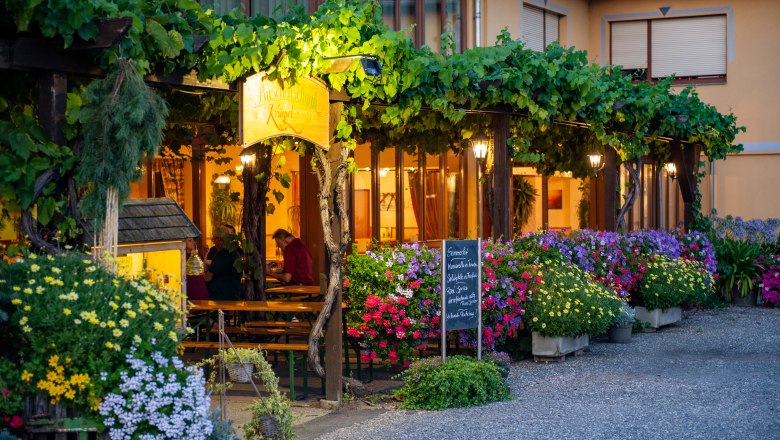 Ein gemütlicher Gastgarten mit blühenden Pflanzen und Weinreben, beleuchtet von warmem Licht. Menschen sitzen an Holztischen unter einem Laubengang.