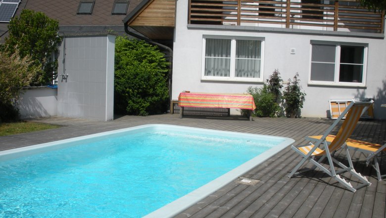 Ein privater Garten mit einem rechteckigen Schwimmbecken, umgeben von Holzterrasse, Liegest&uuml;hlen und einem Haus im Hintergrund.