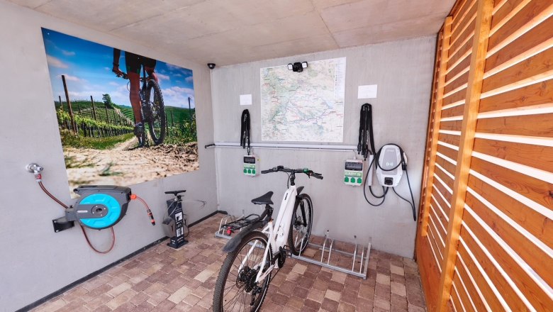 Fahrrad in einer Garage mit Ladestation und Wandbild eines Radfahrers in einem Weinberg.