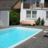 Ein privater Garten mit einem rechteckigen Schwimmbecken, umgeben von Holzterrasse, Liegest&uuml;hlen und einem Haus im Hintergrund.