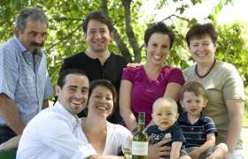 Eine Familie posiert lächelnd im Freien mit einer Flasche Wein auf einem Tisch.