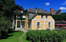 Gelbes Haus mit gr&uuml;nem Garten, im Hintergrund eine Burg auf einem H&uuml;gel.