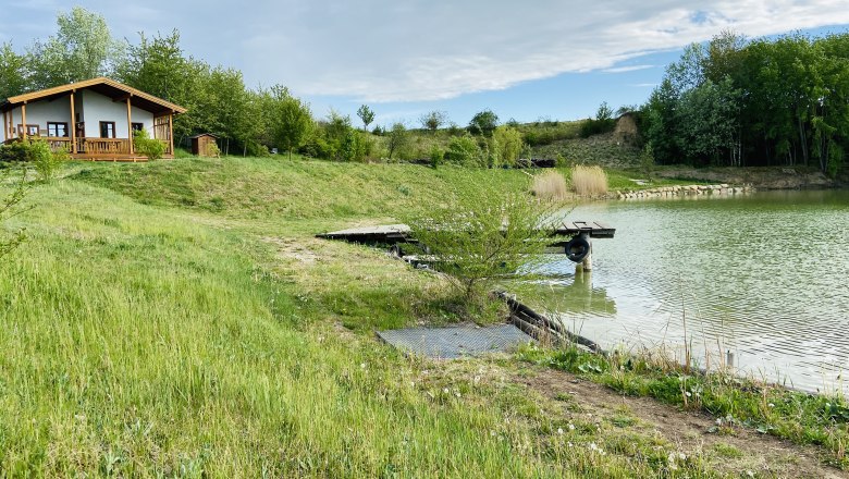 Ein kleines Haus am Ufer eines Teiches mit grüner Wiese und Bäumen im Hintergrund.
