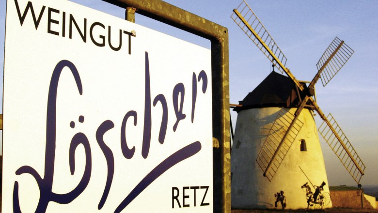 Schild des Weinguts Löscher vor einer Windmühle bei Sonnenuntergang.