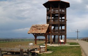 Aussichtsturm aus Holz auf einer Wiese mit Sitzbänken und einem Kreuz im Hintergrund.