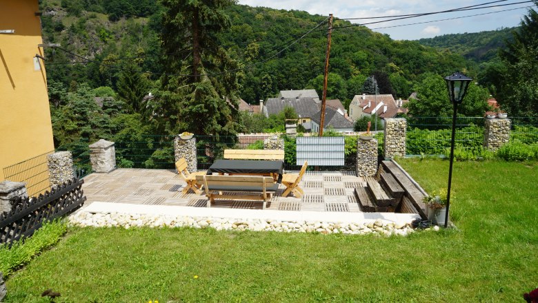 Terrasse mit Holzm&ouml;beln und Blick auf ein Dorf und bewaldete H&uuml;gel.
