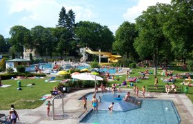 Ein Freibad mit mehreren Pools, einer gelben Wasserrutsche und vielen Menschen, die auf der Wiese entspannen.