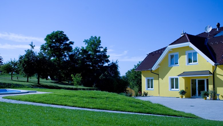 Gelbes Haus mit Garten und B&auml;umen im Hintergrund.